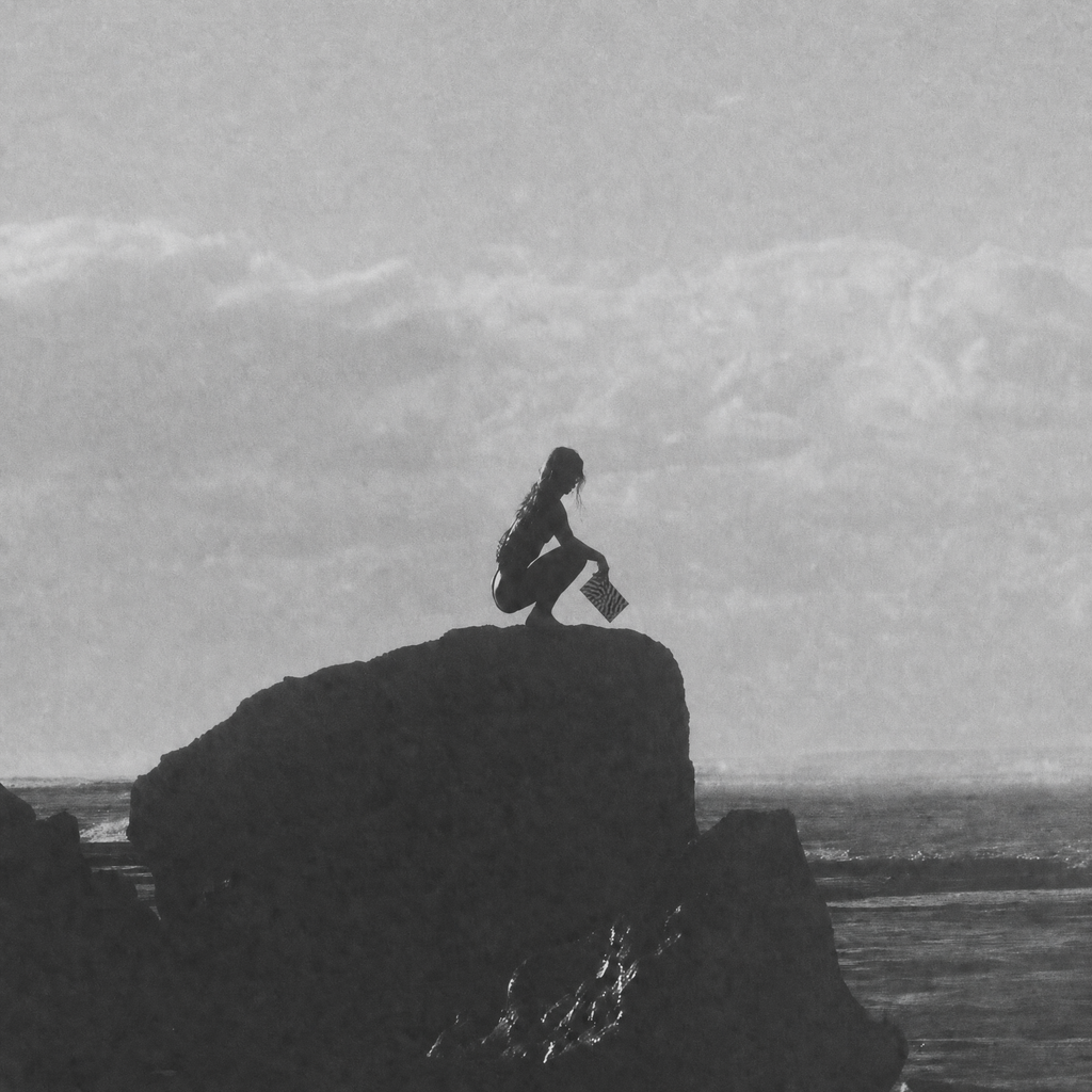 Black and white photograph of a person crouching on a large rock formation over the ocean, with a cloudy sky above.