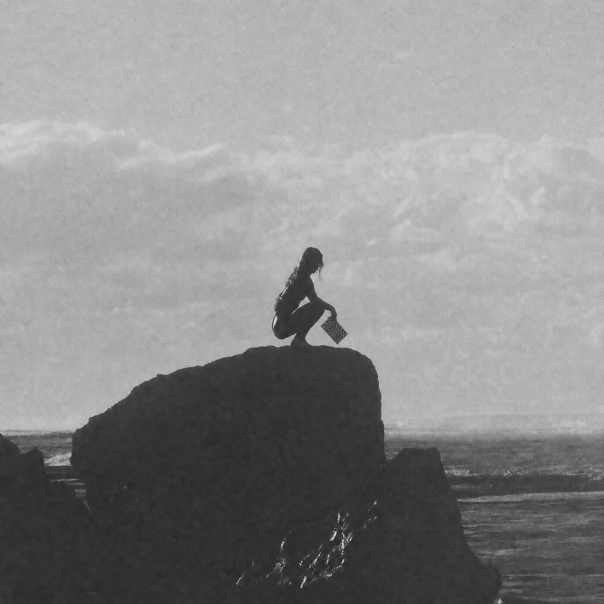 Black and white photograph of a person crouching on a large rock formation over the ocean, with a cloudy sky above.