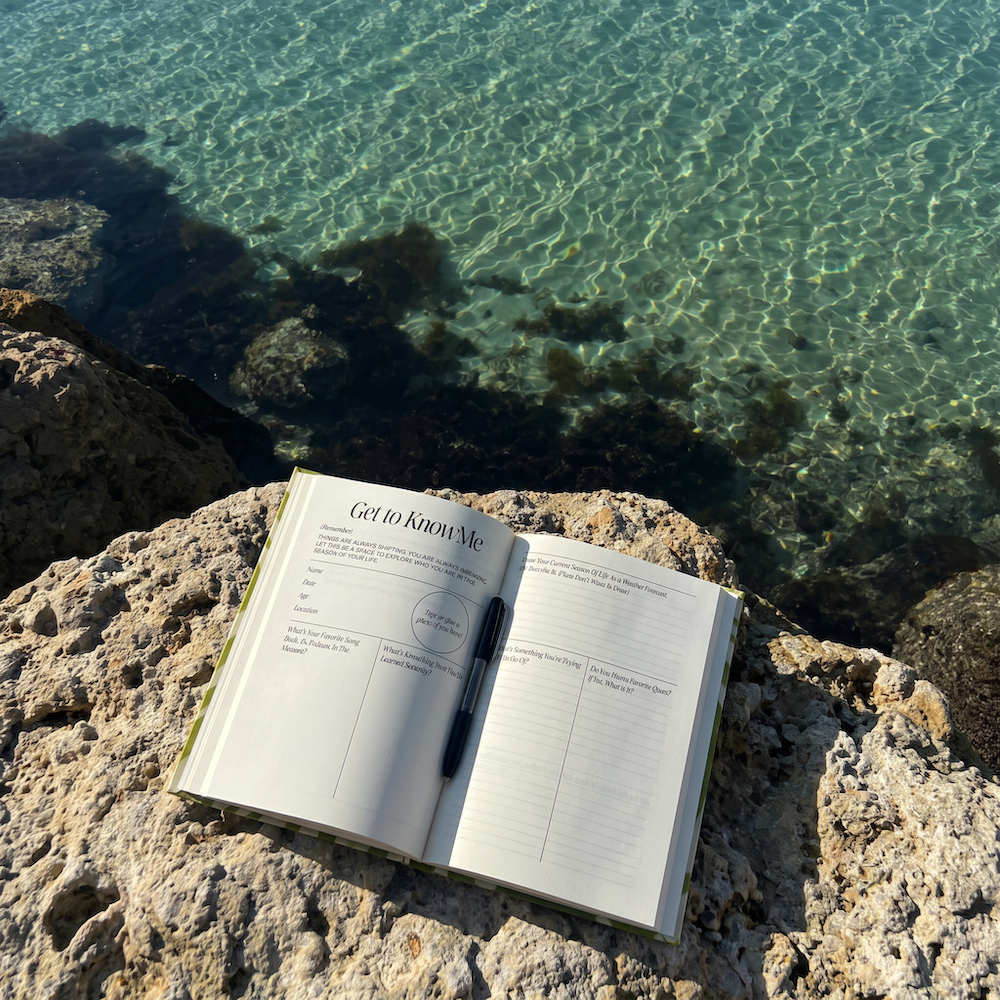 An open notebook with the title 'Get to Know Me' and a black pen resting on it, placed on a rocky shore with clear turquoise water in the background.