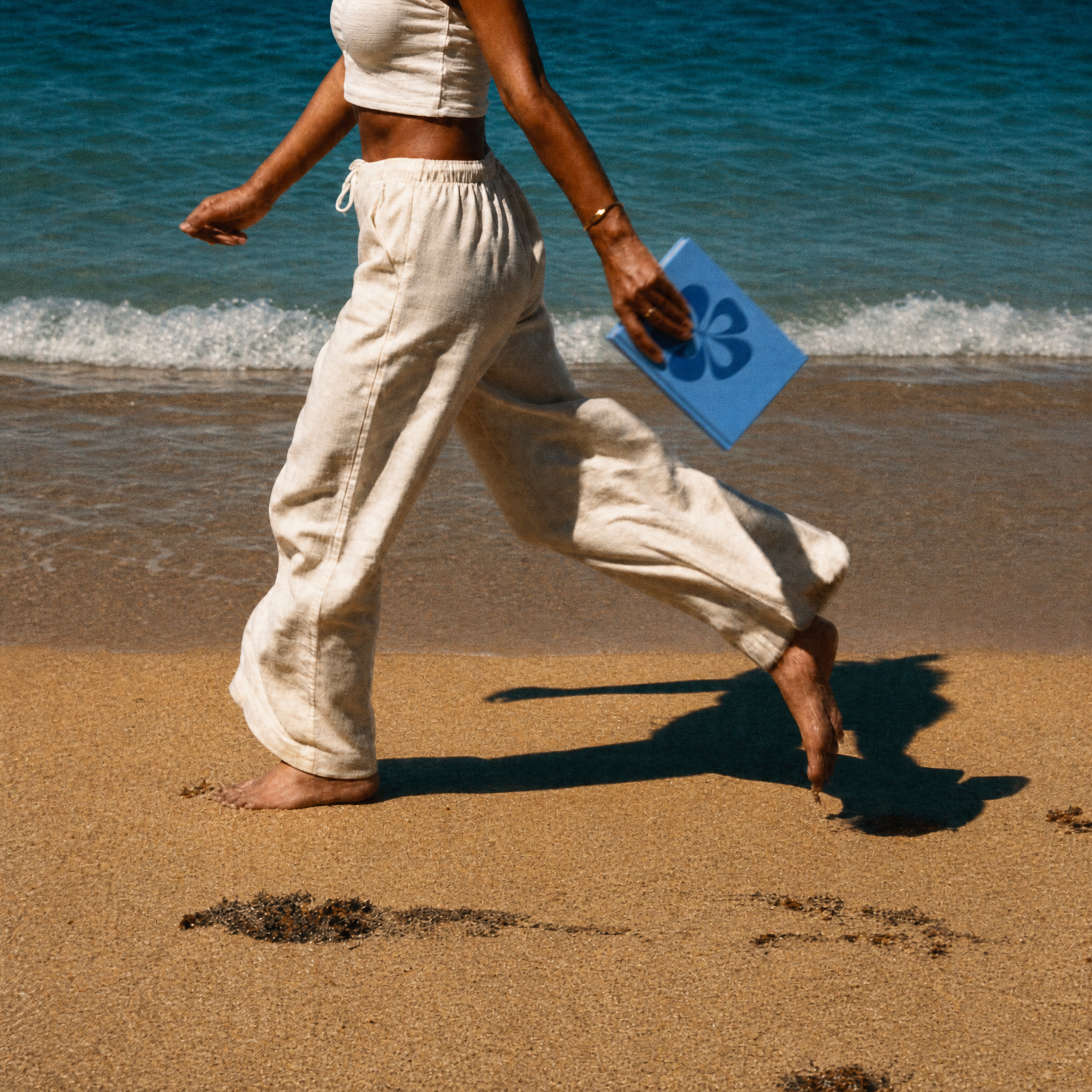 A person walking barefoot on a sandy beach near the ocean, wearing loose off-white cropped pants and a matching top, while holding a blue folder or notebook.