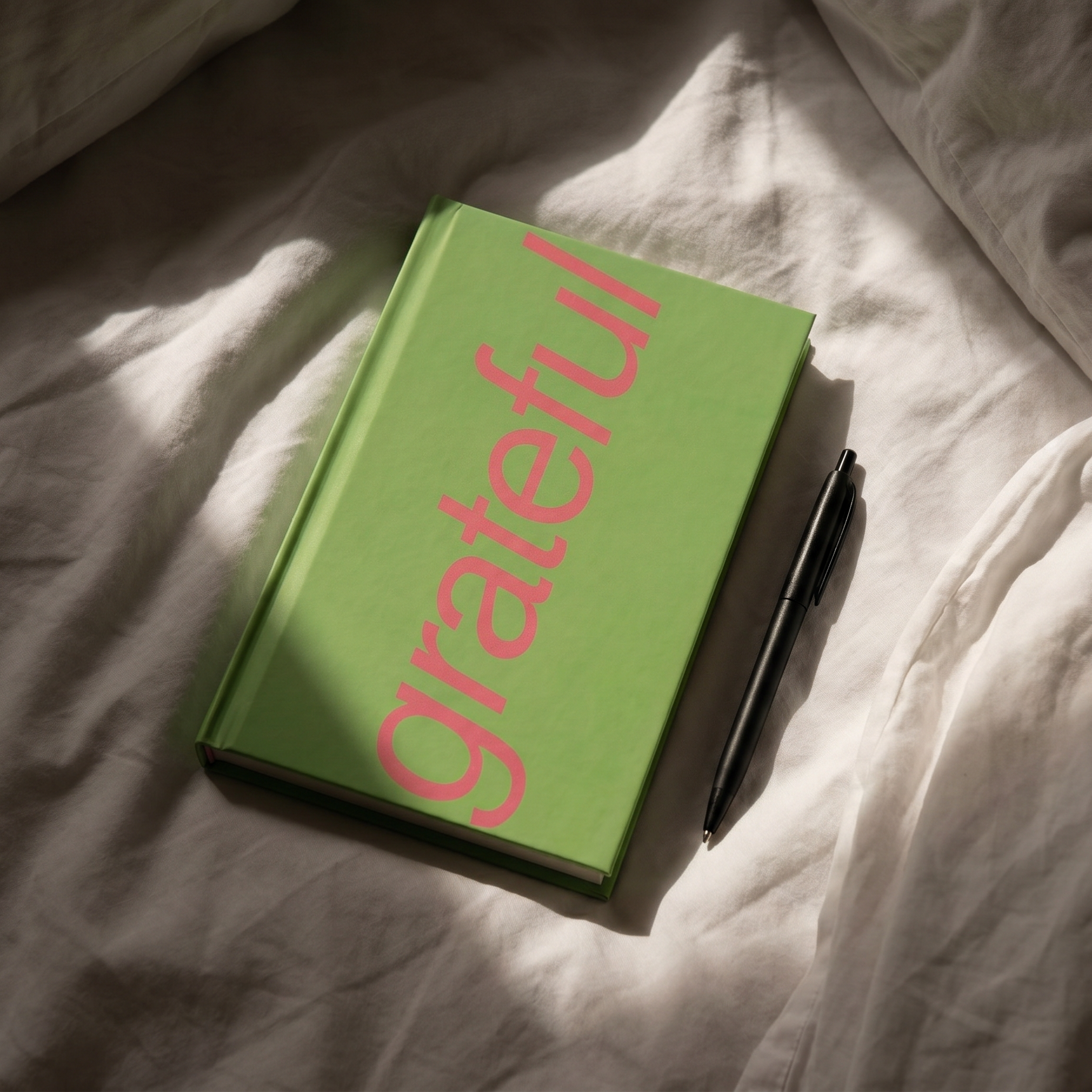 A green hardcover book with the word 'grateful' printed in pink script across the cover, placed on rumpled white bedding with a black pen beside it.