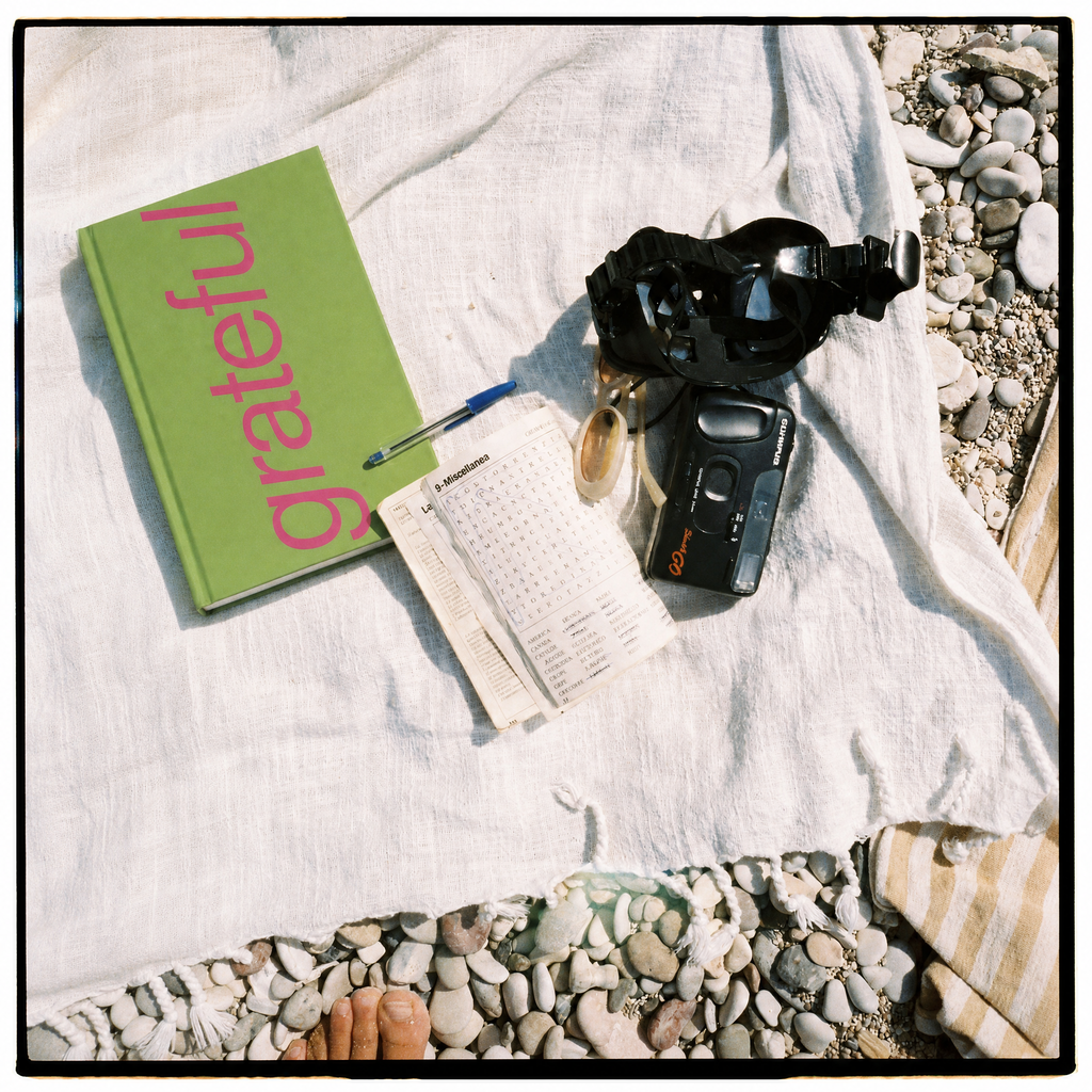 A styled flat-lay on a white linen blanket over pebbles, featuring a green book with the word 'grateful' in pink, a small notebook, a pen, a black camera, and a black headlamp. A foot is visible at the bottom edge.