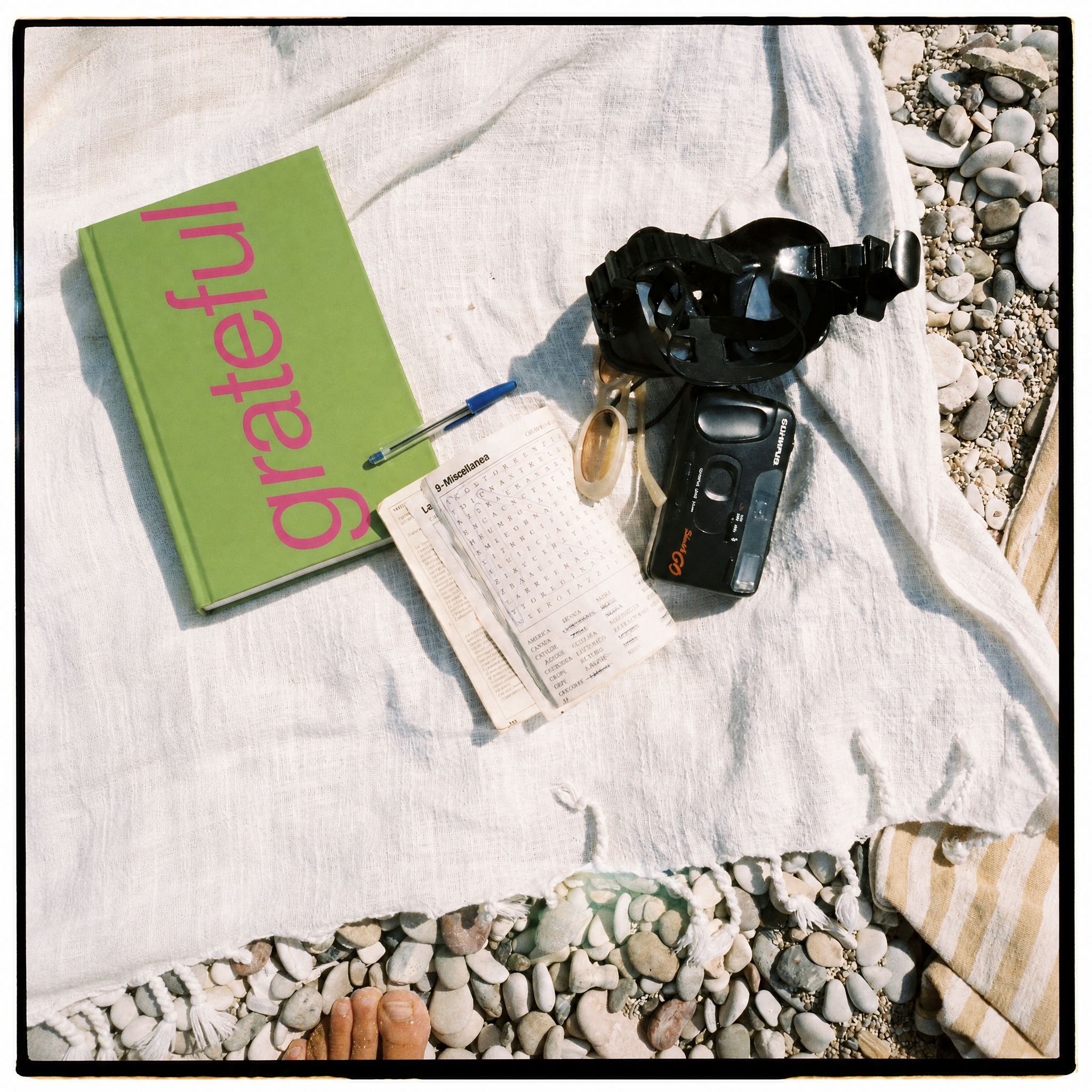 A styled flat-lay on a white linen blanket over pebbles, featuring a green book with the word 'grateful' in pink, a small notebook, a pen, a black camera, and a black headlamp. A foot is visible at the bottom edge.