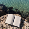 An open lined notebook with a black pen resting on it, placed on a rocky shoreline with clear turquoise water in the background.
