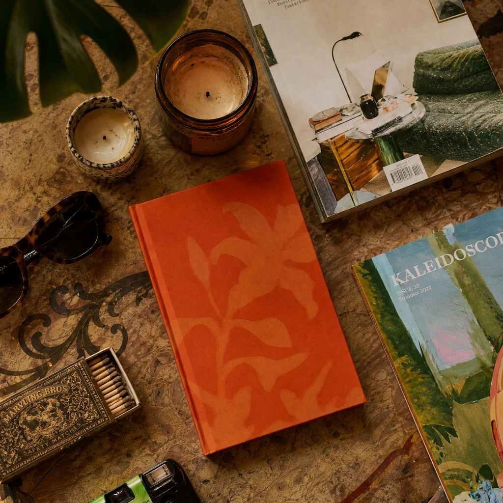 A styled flat-lay featuring a bright orange hardcover notebook with a tonal leaf pattern on the cover, surrounded by a vintage camera, sunglasses, a candle, a matchbox, and two books on a patterned surface.
