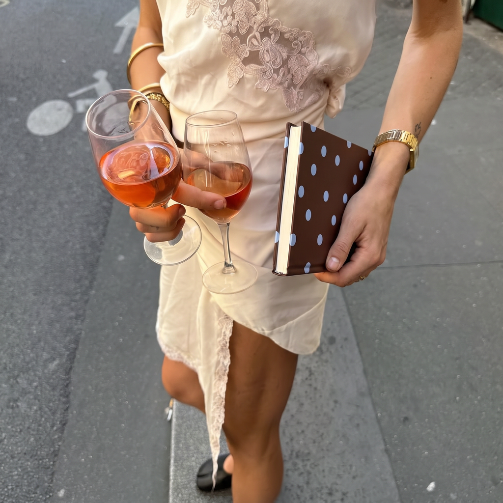 A person outdoors holding two stemmed wine glasses filled with pink drink and a brown notebook with white polka dots, wearing a cream lace-trim dress and gold jewelry.