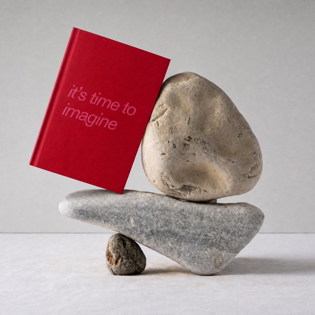 A red hardcover book with the text 'it's time to imagine' in white, balanced on top of stacked gray and beige stones against a light gray background.