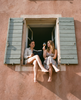 Two women sitting in an open window, one reading a book and the other holding a glass, against a pink wall.