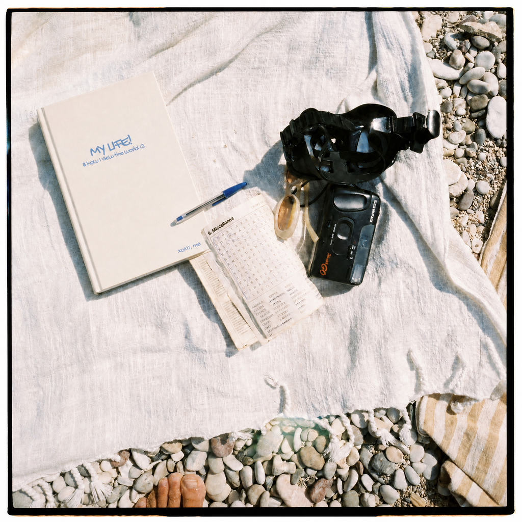 A styled flat-lay of a white notebook with the text 'MY LIFE! A Memoário dos Análisis' and a pen, alongside a small open notebook, a clear bottle, and a black action camera, arranged on a white linen cloth over a bed of pebbles.