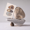 A square art print with minimalist abstract stone illustrations in earthy tones, propped up against a stack of three natural river stones on a light gray surface against a neutral gray background.