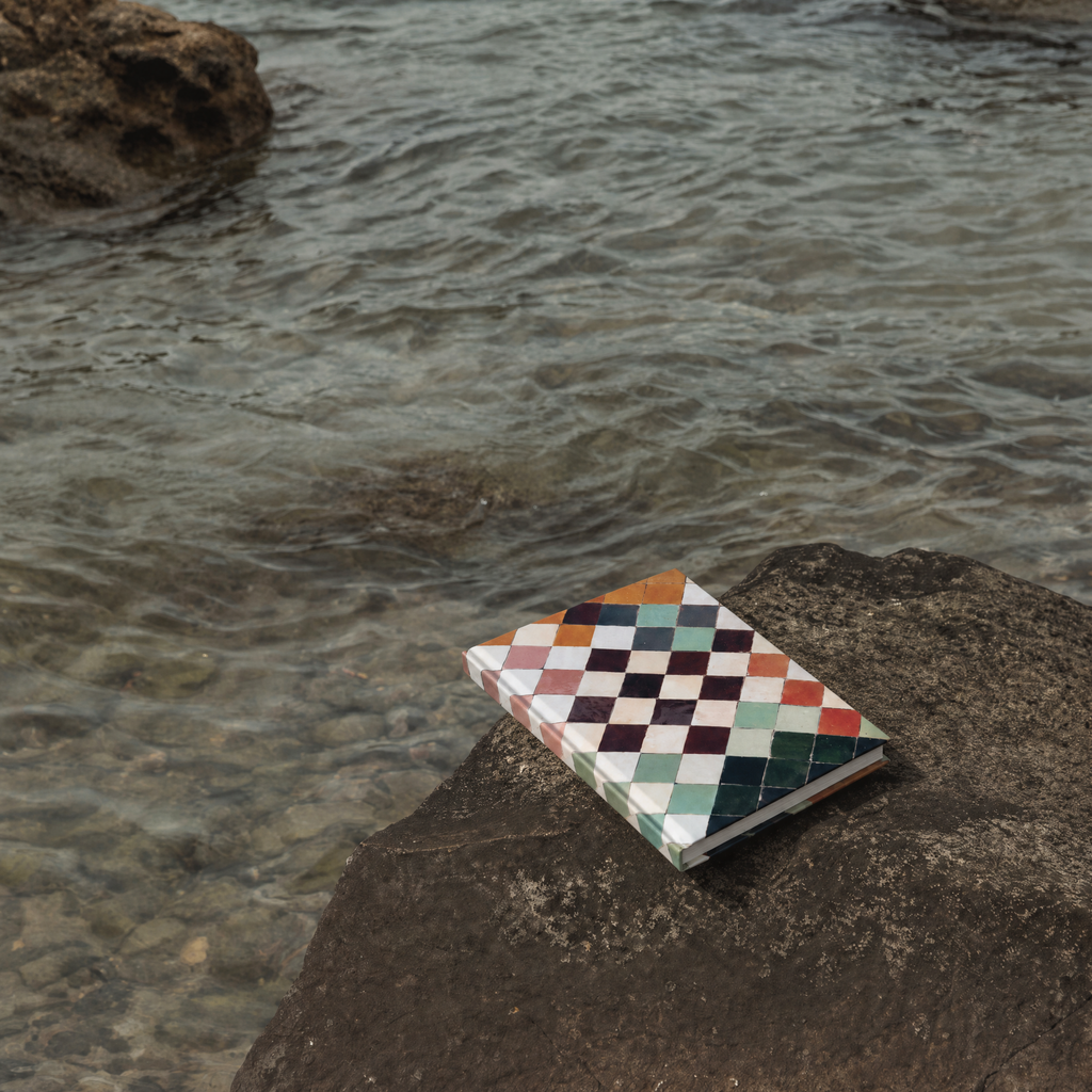 A hardcover notebook with a colorful geometric checkered pattern lies on a dark rock next to a body of water.