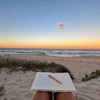 An open notebook with a wooden pen rests on a person's lap on a sandy beach, with the ocean and a sunset sky in the background.