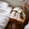 A styled bedroom scene with a wooden bedside table next to a bed with white linens. On the table are an open book, a closed book with abstract oval illustrations on the cover, a green ceramic mug, a small card, and a glass jar holding dried pink and beige flowers.